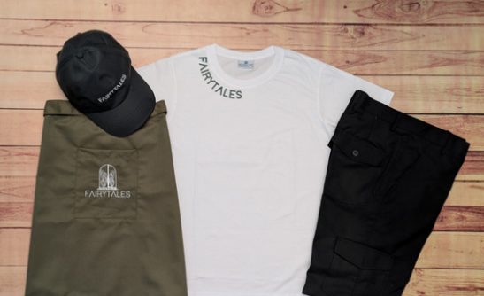 Complete cafe uniform flatlay, white t-shirt, black cap, olive apron and black cargo trousers embroidered 'Fairytales'